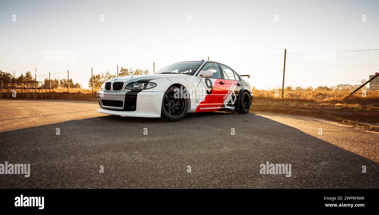 white-red-black BMW E46 racecar near the fence and dry grass. Three quarter front view of widebody sedan at sunset. Stock Photo