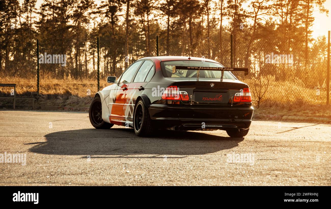 white-red-black BMW E46 racecar near the fence and dry grass. Three quarter rear view of widebody sedan at sunset. Stock Photo