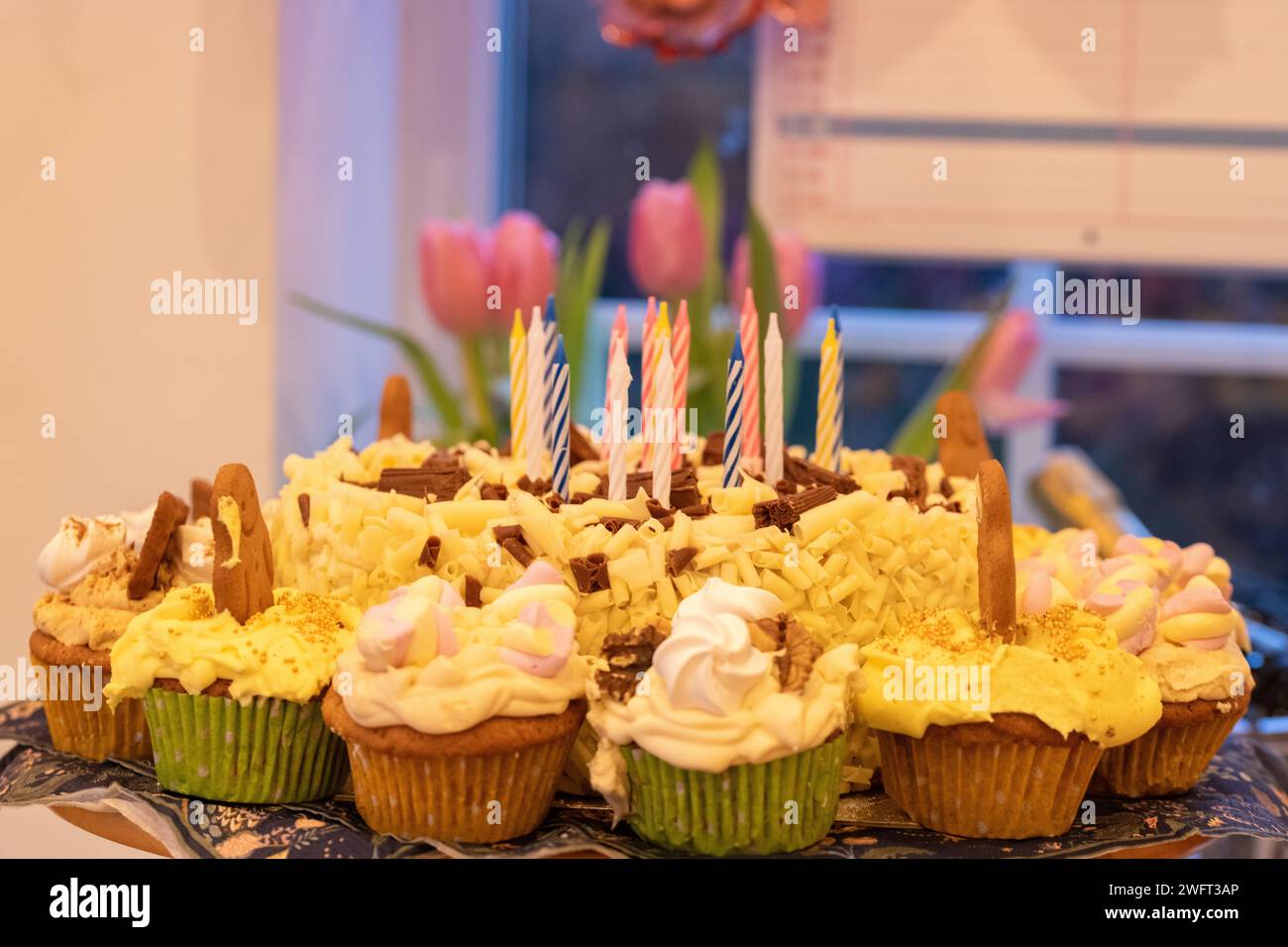 A white chocolate birthday cake with unlit candles surrounded by homemade cupcakes covered in individual decoration and butter icing Stock Photo
