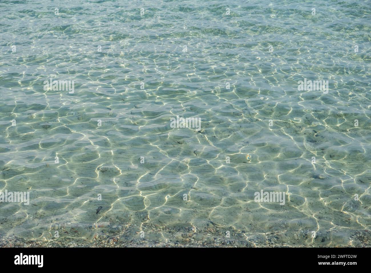 Clear shallow light blue sea, ripple transparent sparkle water surface background texture. Underwater pebble and sand, destination Greece. Space Stock Photo
