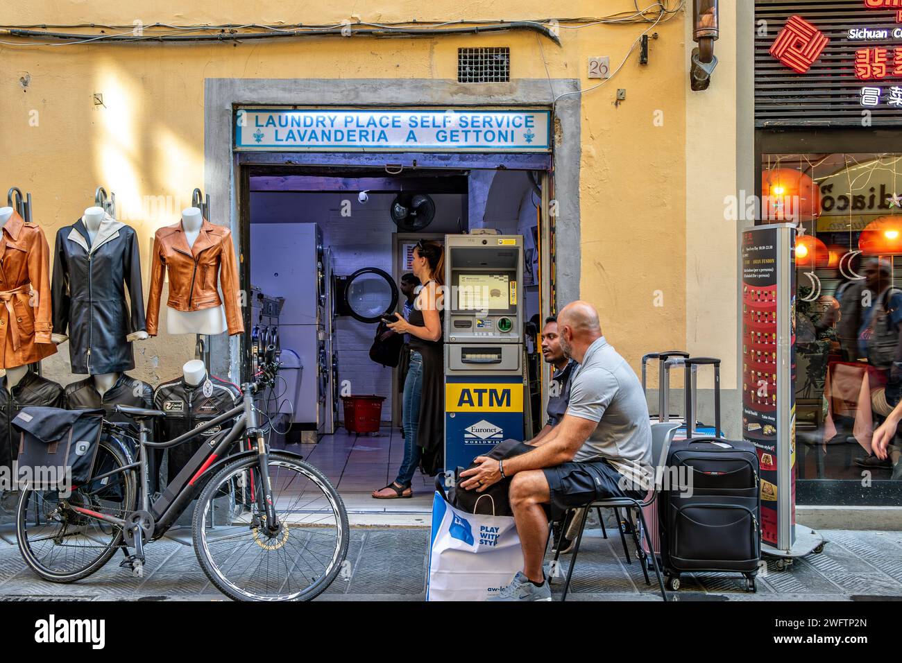 A woman using a launderette, lavanderia a gettoni, with two men sitting down outside waiting on Via Faenza, a street in Florence,Italy Stock Photo