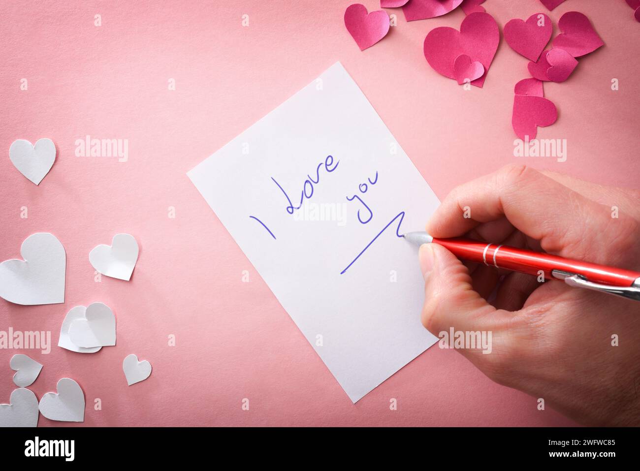 Hand writing I love you on white sheet on pink background with pink and white paper heart cutouts. Top view. Stock Photo