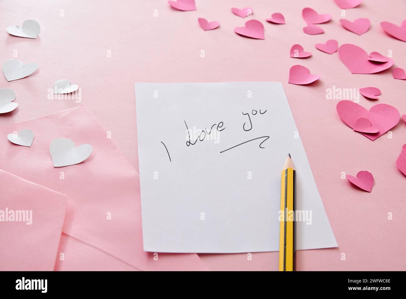 Note with writing I love you on white paper on a pink background full of heart-shaped cutouts. Top elevated view. Stock Photo