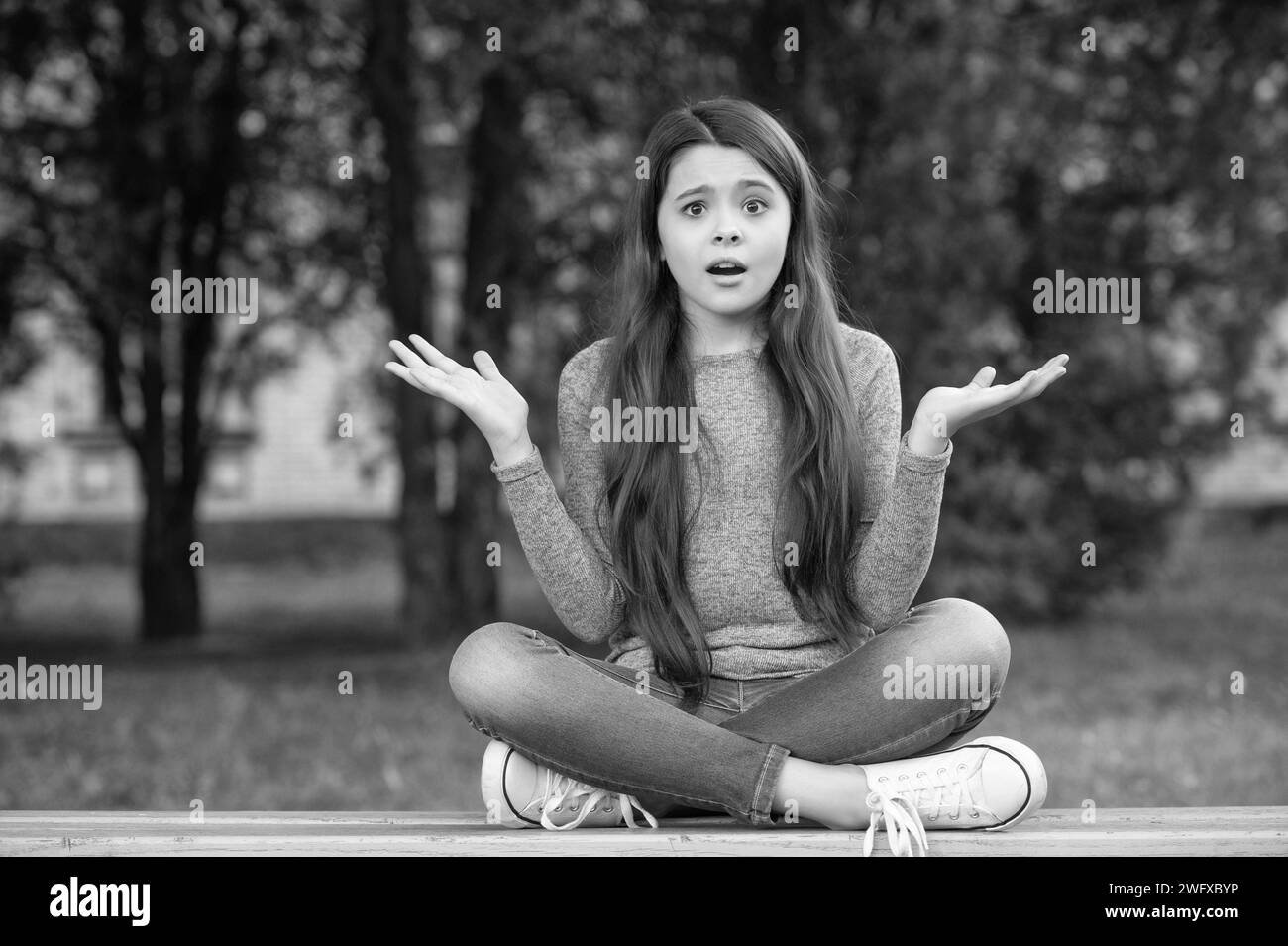 Shocked teenage girl shrugging shoulders sitting legs crossed on bench outdoors, shock. Teenage girl being in shock. Stock Photo