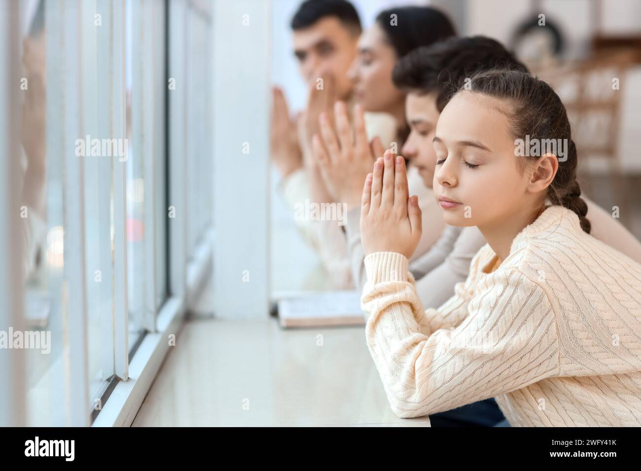 Little girl praying with her family near window at home, closeup Stock Photo