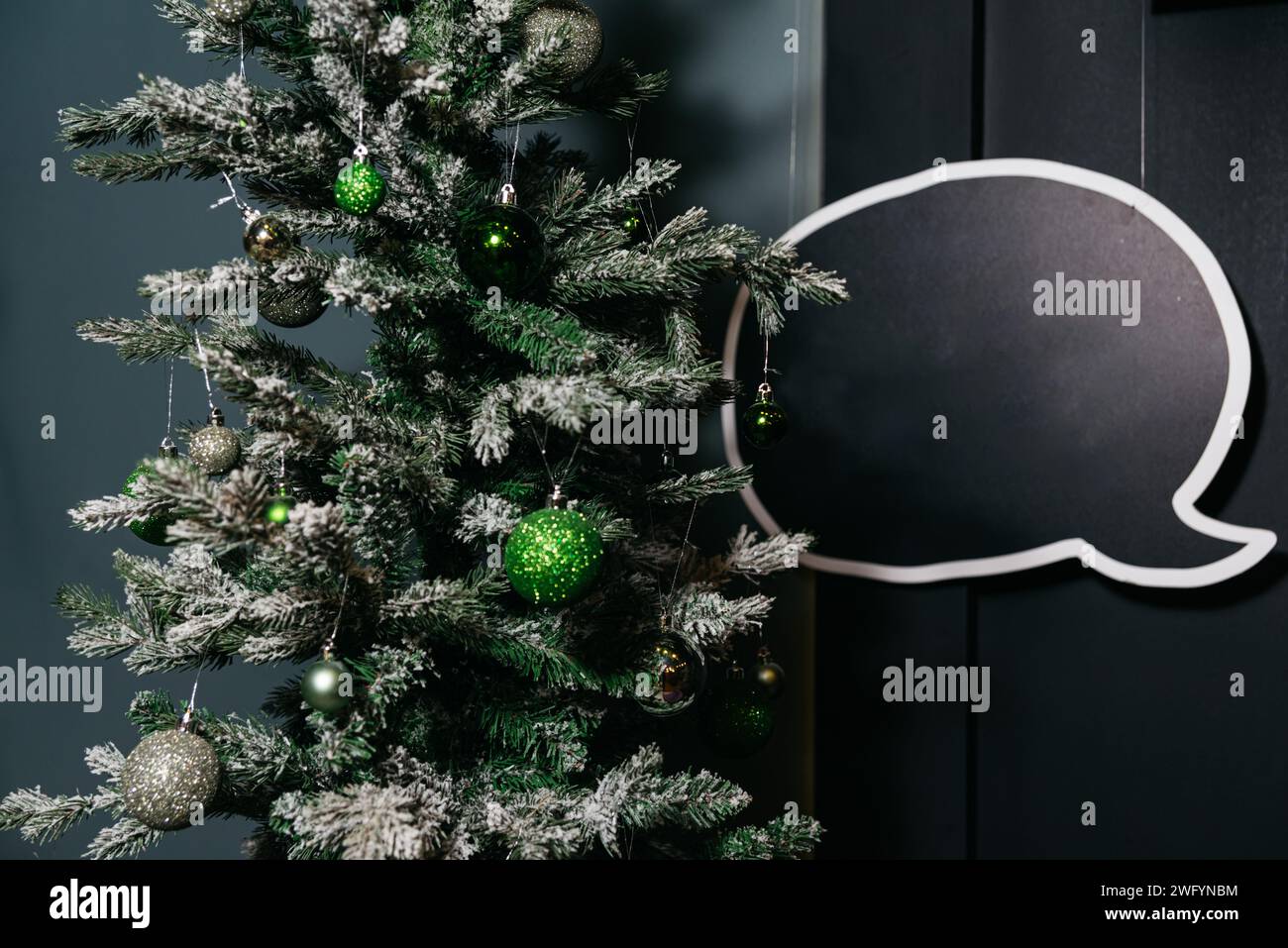 A frosted Christmas tree adorned with green ornaments and a blank speech bubble decoration, set against a dark backdrop. Stock Photo