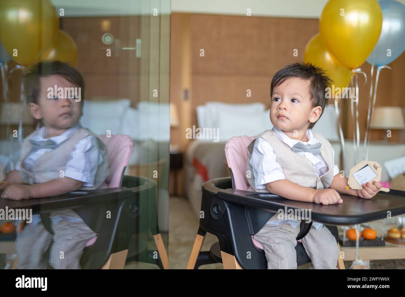 Cute one year old multiracial baby boy celebrating his first birthday with balloons sitting in a high feeding chair at home. He is dressed in a beige Stock Photo