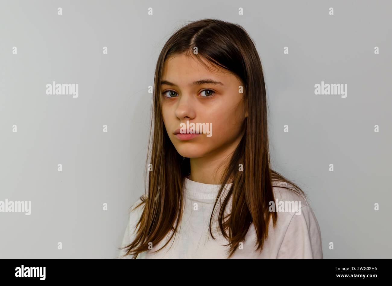 A teenage girl with long hair wearing a white t-shirt looks at the camera against a light colored background. Stock Photo