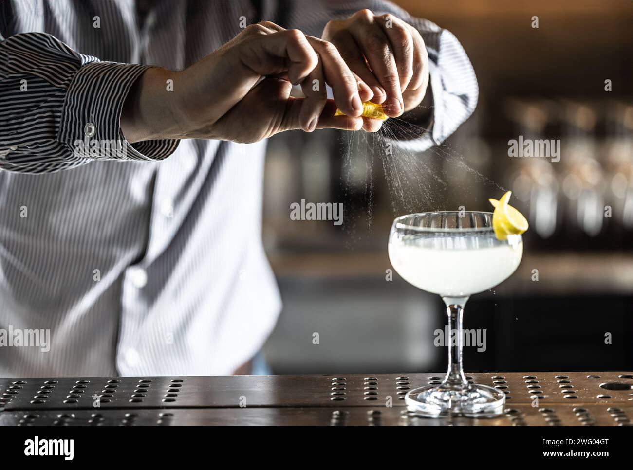 A bartender squeezes a lemon peel and squirts the juice onto a French 75 cocktail drink on the bar counter. Stock Photo