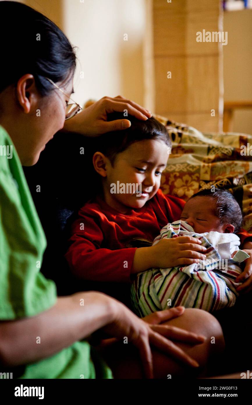 A little boy holds his newborn baby brother, while his mother looks on