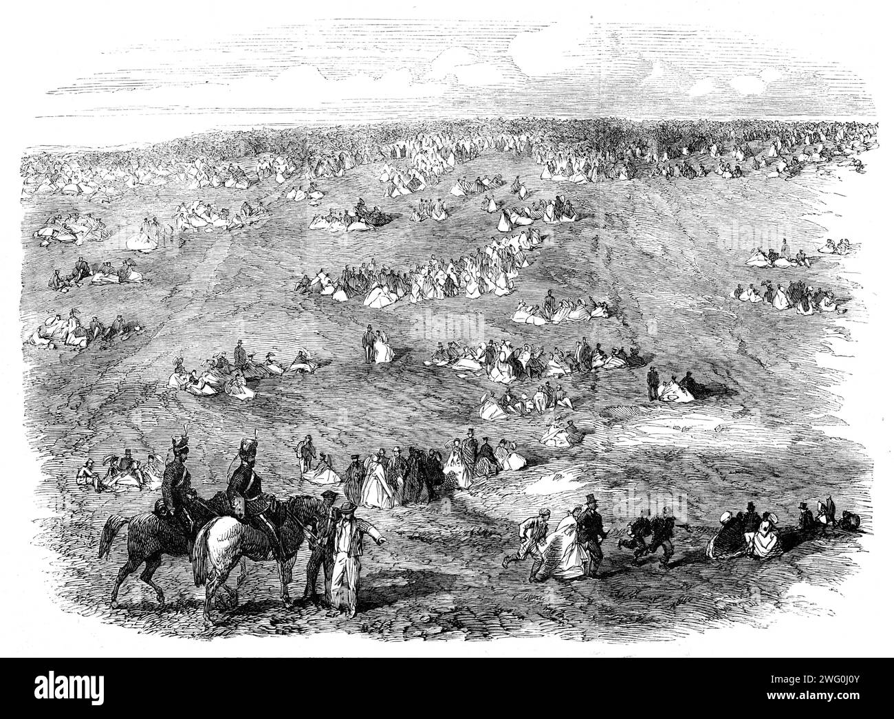 The Volunteer Field-Day at Brighton: people on the Downs, 1862. 'Nothing could be finer than the approach of something like 12,000 men in close column, at quick march, up the steep hill leading to the racecourse...The whole aspect of the field, as seen from the Grand Stand, was magnificent in the extreme...On each side, as far as the racecourse extended, was a dense body of spectators, which appeared to stretch away for miles...a few artillery troops could by the aid of a good glass be seen unlimbering two or three guns...the furthermost brigade [offered], in the bright scarlet uniform of two Stock Photo
