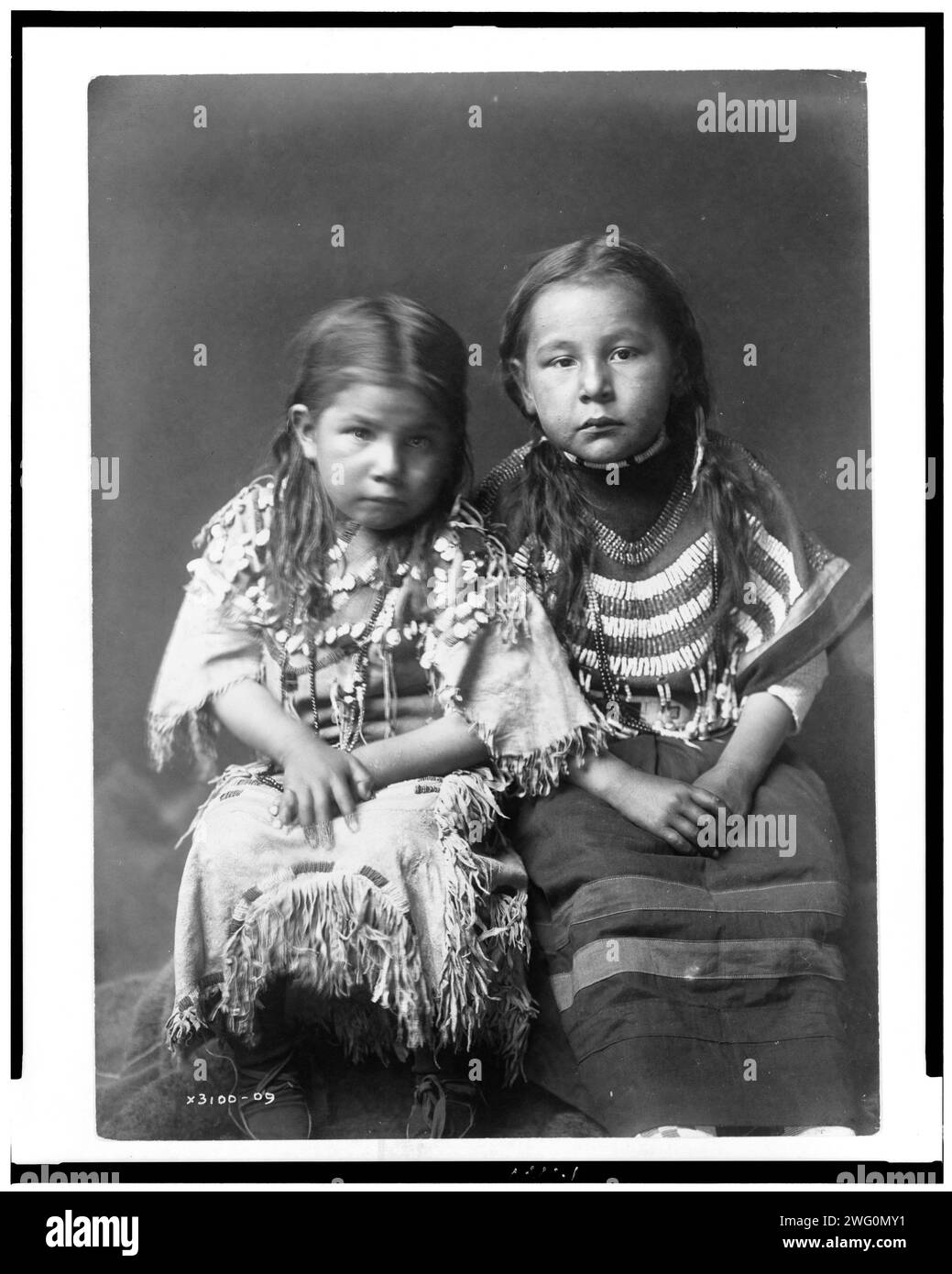 Bull Shoe's children, c1910. Two girls, full-length, seated, facing front, with loose hair, one wearing buckskin dress decorated with elks' teeth, one wearing beaded cloth dress. Stock Photo