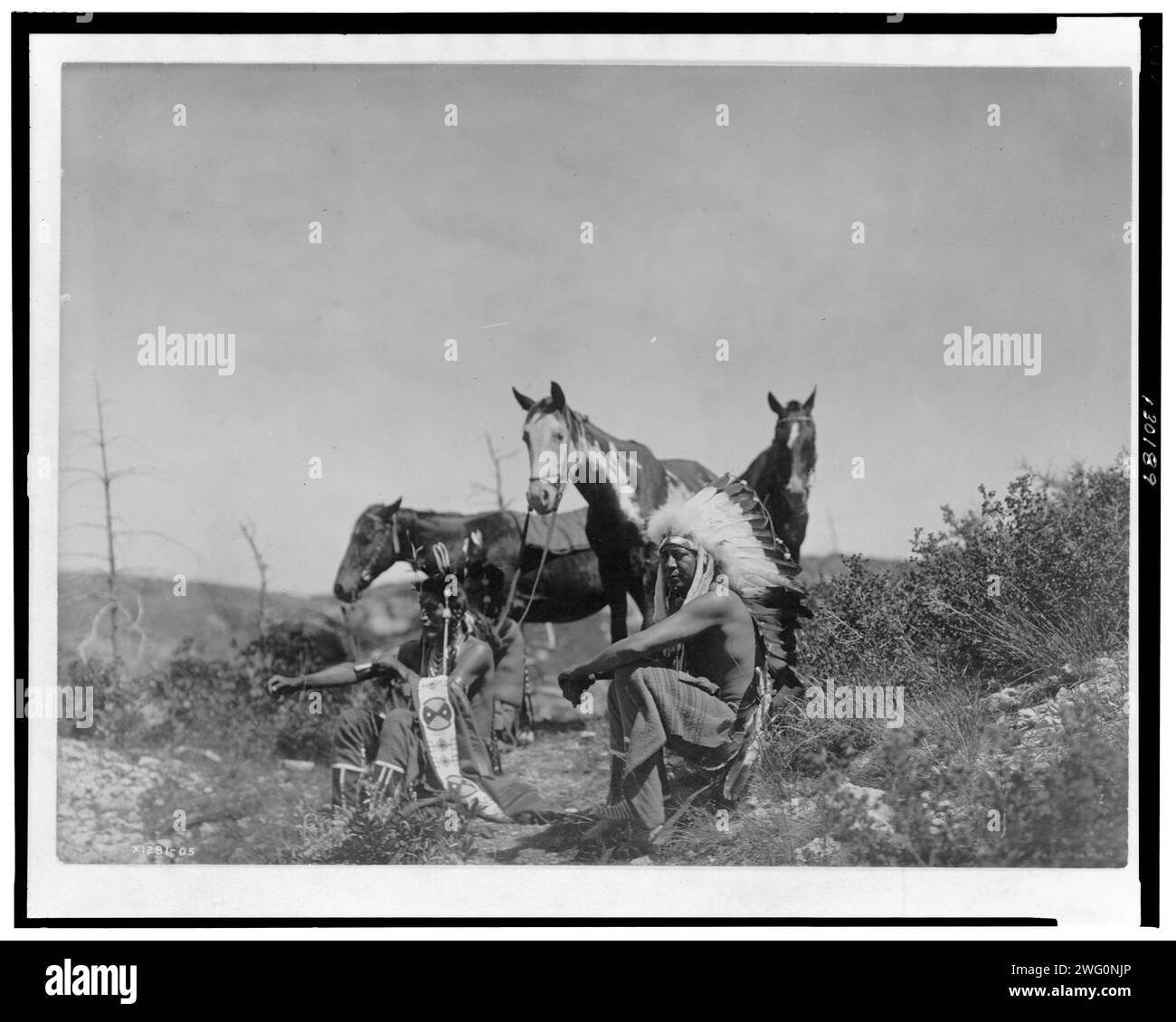 The Talk, c1905. Three Crow men, seated in rocky area with low growing shrubs, their horses rest nearby. Stock Photo
