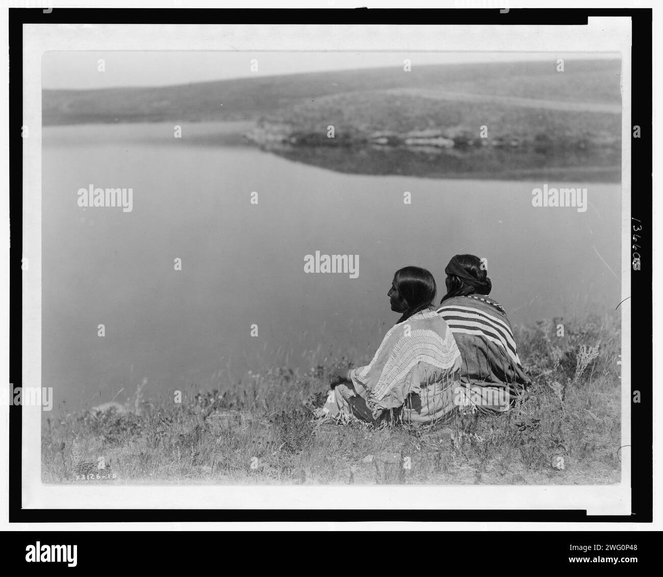 An idle hour, Piegan, c1910. Photograph shows two Piegan Indians sitting on grassy area above a body of water. Stock Photo