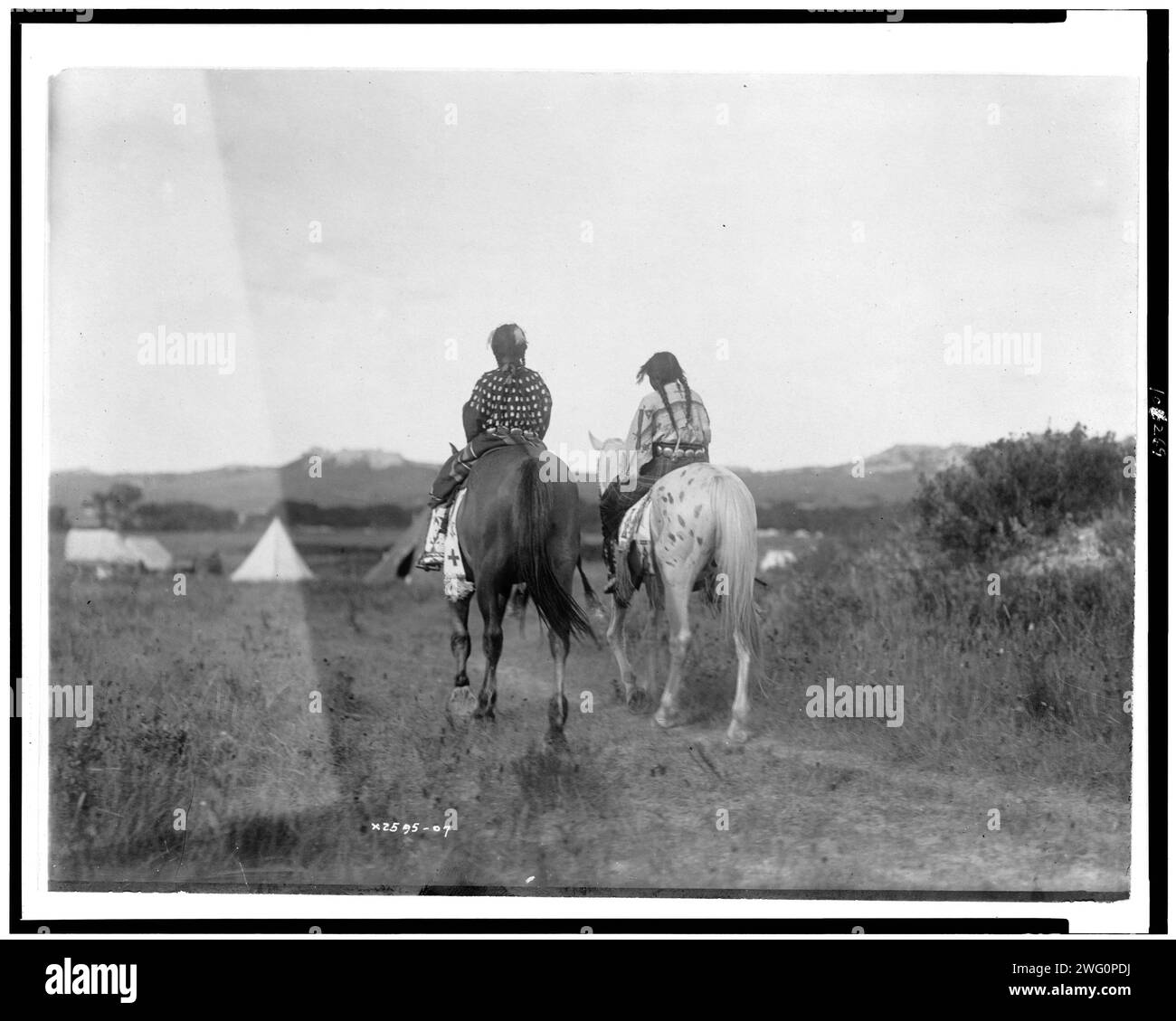 Two daughters of a chief on horseback, riding away from camera toward tents in background, c1907. Stock Photo