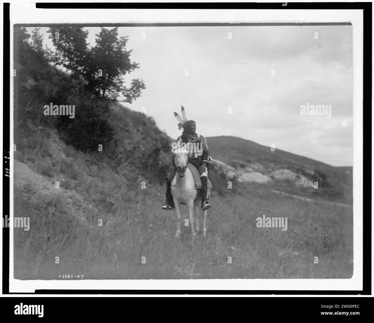 Sioux Indian on horseback, wearing two feathers, beaded buckskin shirt, and leggings, with hills in background, c1907. Stock Photo