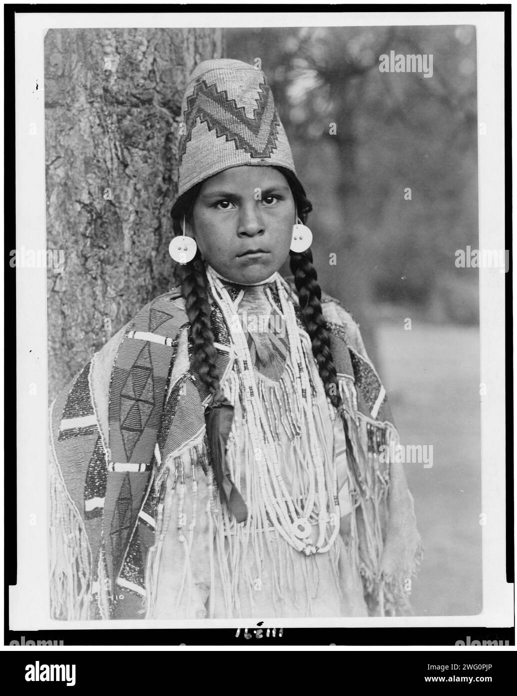 Umatilla maiden, c1910. Umatilla woman, half-length portrait, facing front, standing in front of tree, wearing beaded buckskin dress, shell bead necklaces, shell disk earrings, and woven grass hat. Stock Photo