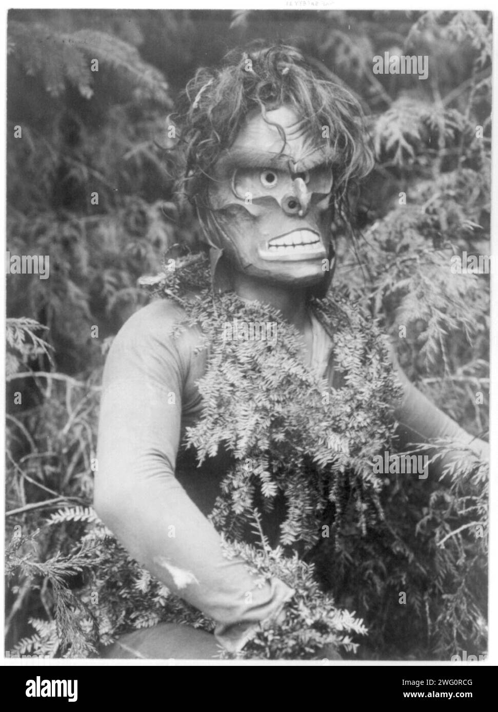 Paqusilahl-Qagyuhl, c1914. Dancer representing Paqusilahl (&quot;man of the ground embodiment&quot;), wearing a mask and shirt covered with hemlock boughs, representing paqus, a wild man of the woods. Stock Photo