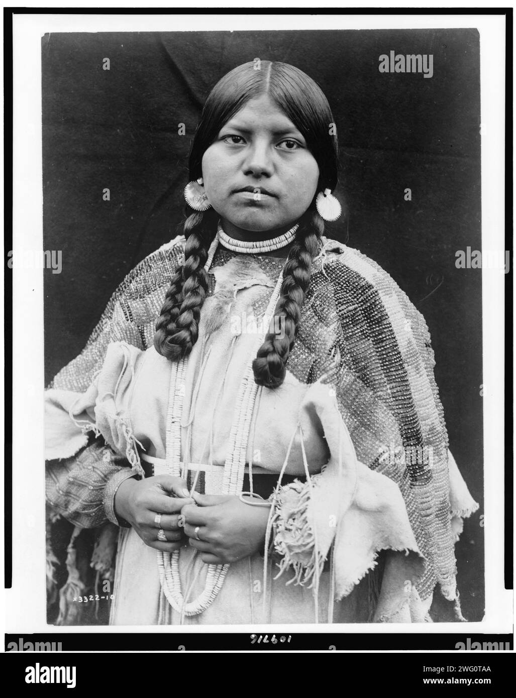 Cayuse woman, half-length portrait, standing, facing front, braids, shell disk earrings, shell bead choker and shell beads around neck, beaded buckskin dress with beaded belt, c1910. Stock Photo