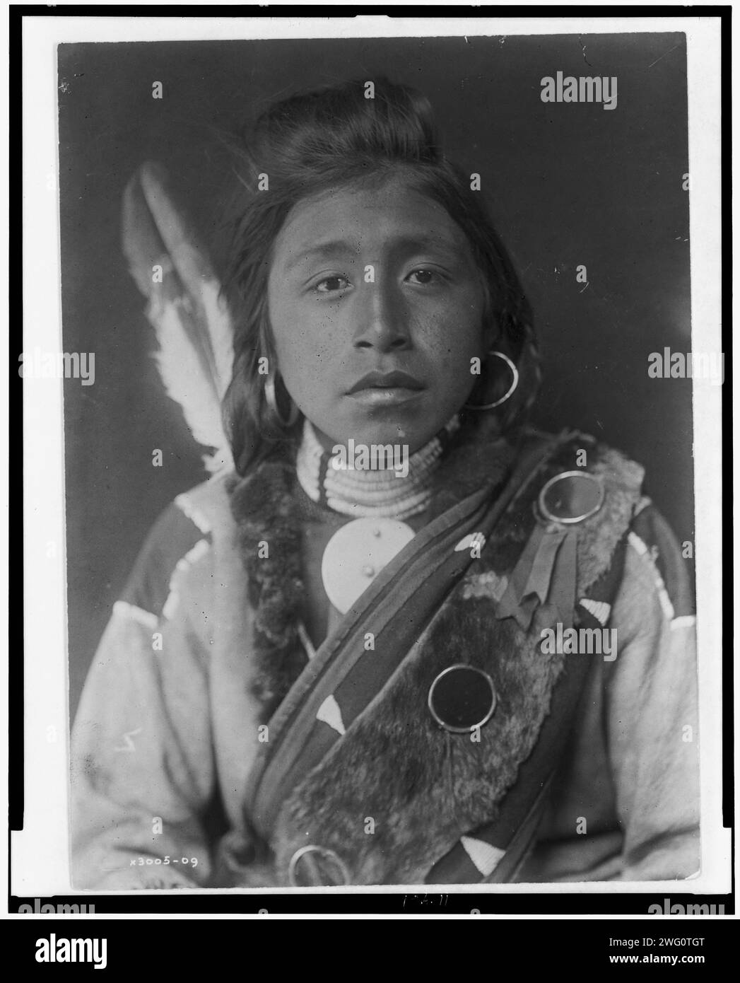 Kashhila-Wishham. Male Chinookan Indian, head-and-shoulders portrait, facing front, with pompadour, fur wrapped braids, bead choker around neck, beaded buckskin shirt, fur sash over left shoulder, c1910. Stock Photo