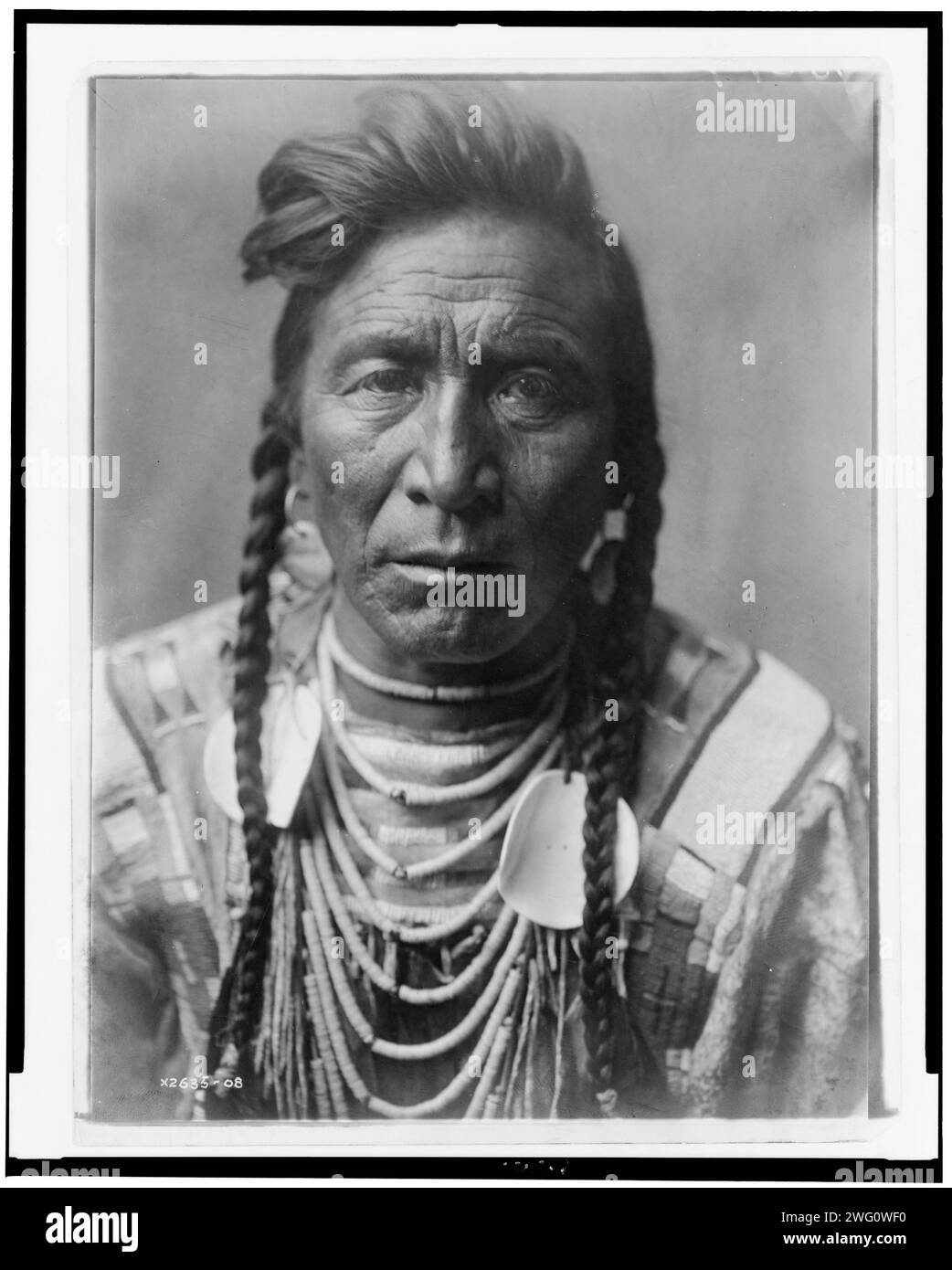 Strike On his Head, Crow Indian, Montana, head-and-shoulders portrait, facing front, loose forelock, braids, beaded buckskin shirt, beads and two large shell disks around neck, c1908. Stock Photo