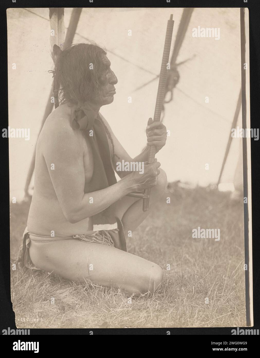 Slow Bull, 1907. Photograph shows three-quarter portrait of Slow Bull inside tipi, facing right, sitting in grass holding a pipe. Stock Photo