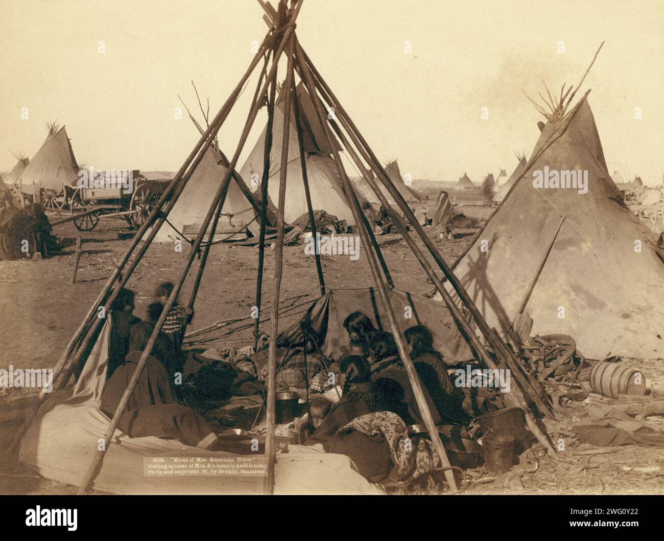 Home of Mrs American Horse Visiting squaws at Mrs A's home in hostile camp, 1891. Oglala women and children seated inside an uncovered tipi frame in an encampment, most are looking away from the camera, probably on or near Pine Ridge Reservation. Stock Photo