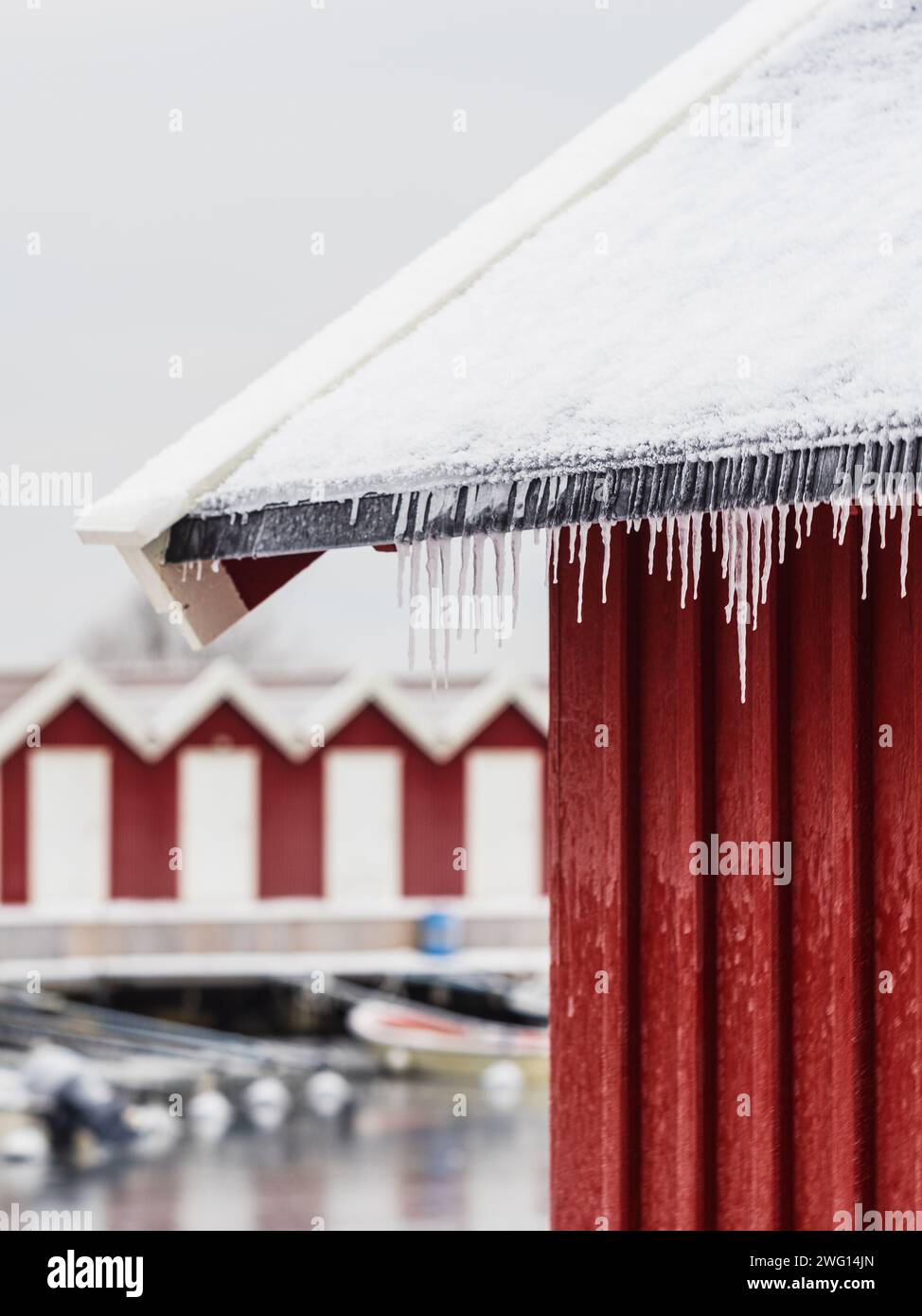 Icicles glisten as they hang from the edge of traditional red cabins beside a tranquil, snow-covered dock. Stock Photo