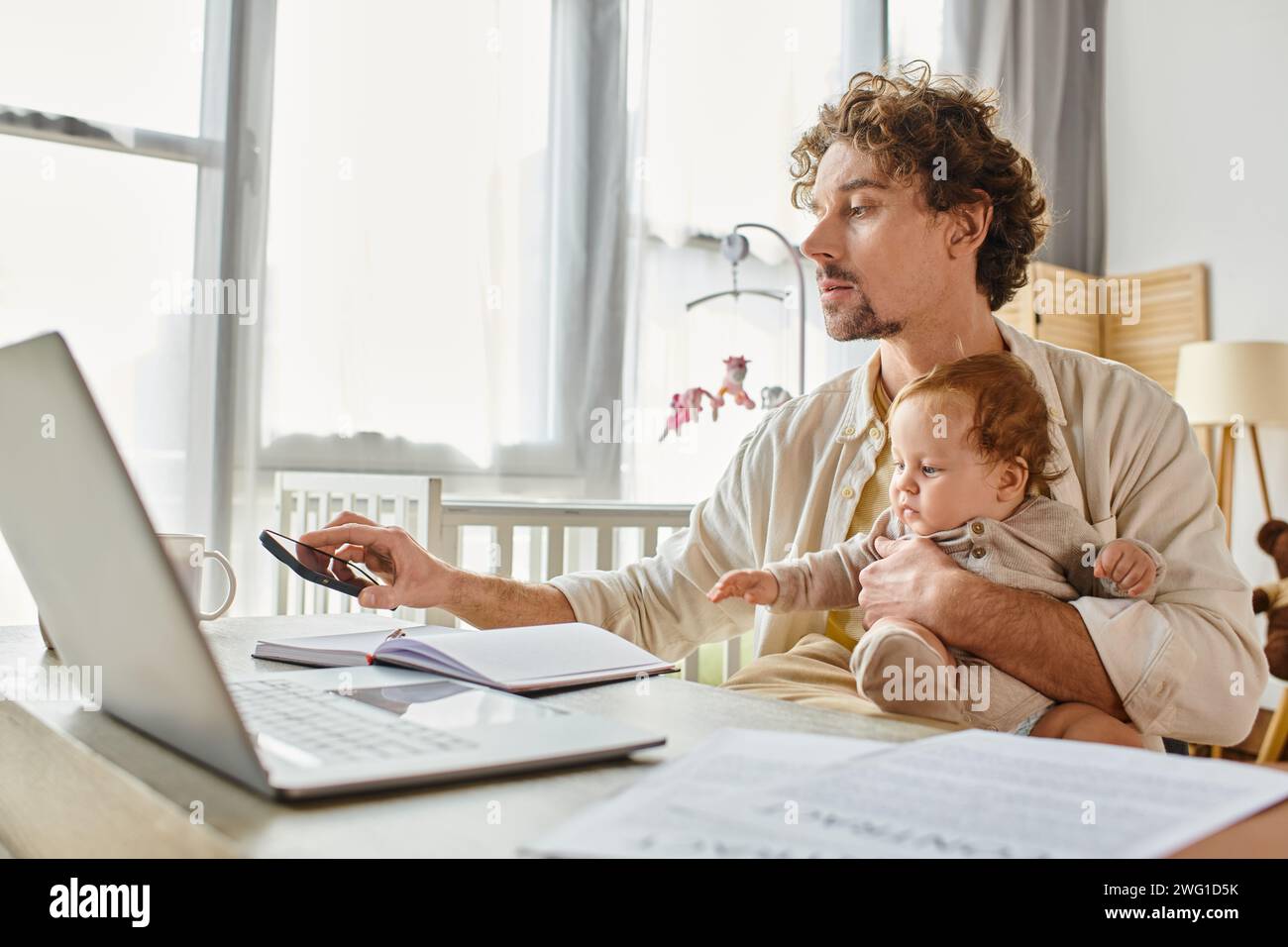 father holding his baby son while taking his smartphone and working ...