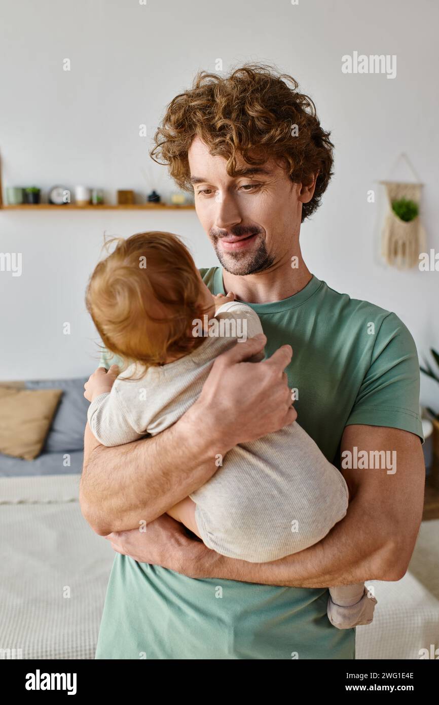 caring father with curly hair and beard holding in arms his infant boy in baby clothes, parenting Stock Photo