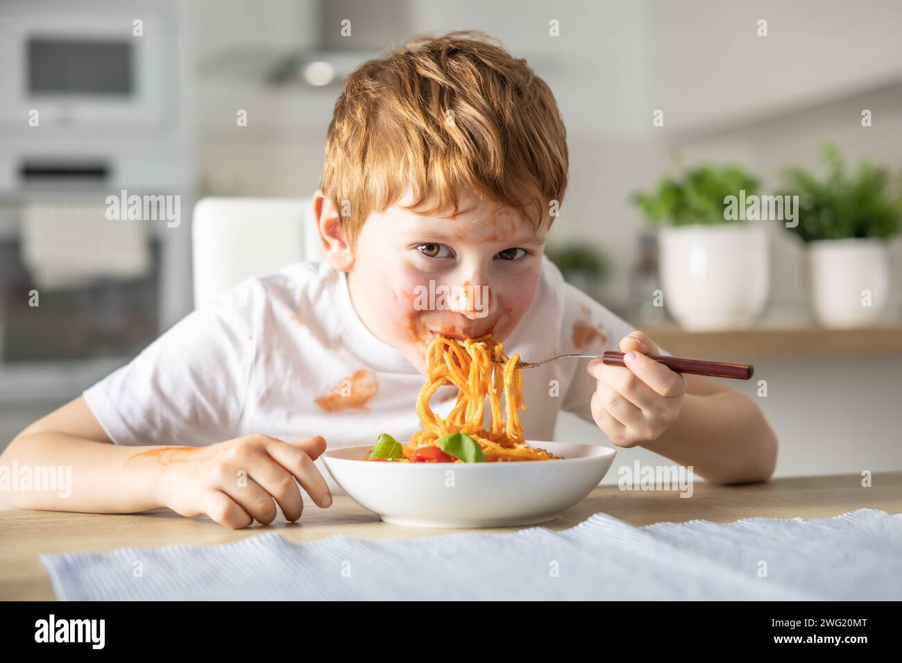 A cute little boy is eating spaghetti bolognese for lunch in the kitchen at home and is covered in ketchup. Stock Photo