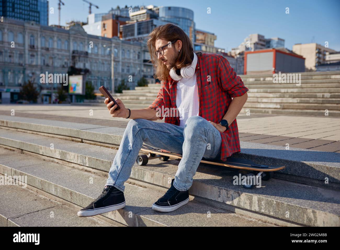 Hipster guy with longboard having rest while sitting on steps and using ...
