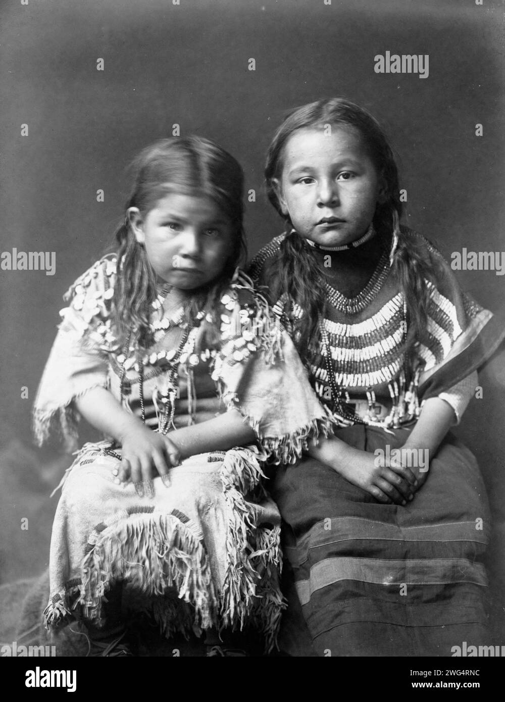 Bull Shoe's children, c1910. Two girls, full-length, seated, facing front, with loose hair, one wearing buckskin dress decorated with elks' teeth, one wearing beaded cloth dress. Stock Photo