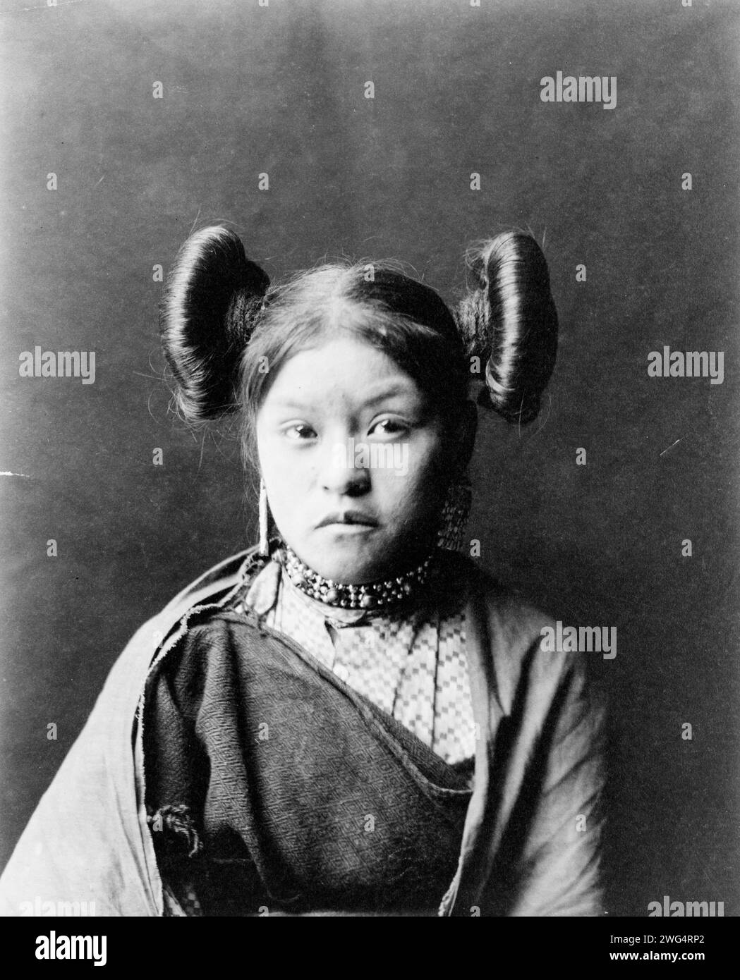 Gobuguoy, Walpi girl, half-length portrait, facing front, hair tied in swirls on sides of head, metal bead and bell choker, printed cotton dress, cotton shawl around shoulders, c1900. Stock Photo
