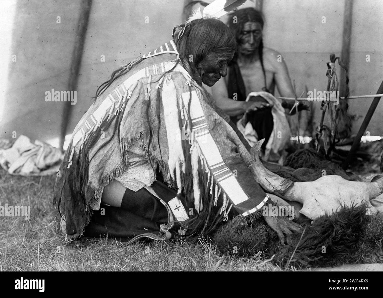 Hu Kalowa Pi-Removes the covering, c1907. Interior of tepee, man kneeling on ground remouving buffalo hide around skull on ground, another male behind altar warming hands by fire. Stock Photo