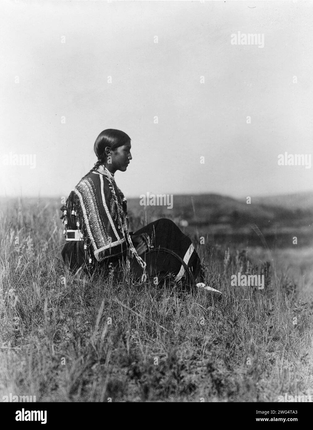 Day dreams-Piegan, c1910. Young Piegan Indian woman, wearing beaded dress, seated on hill. Stock Photo