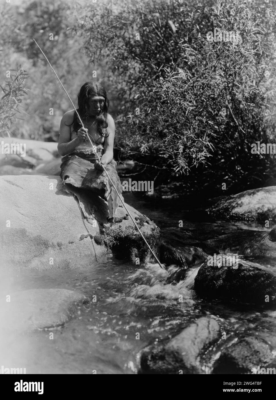 On the Merced-Southern Miwok, c1924. Miwok man holding spear, sitting on boulder in a creek. Stock Photo