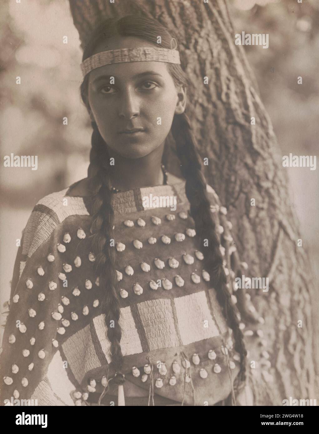 Lucille [B], c1907. Photograph shows Lucille, a Native woman, half-length portrait, standing next to tree, facing front, wearing a headband and seashell decorated buckskin dress. Stock Photo