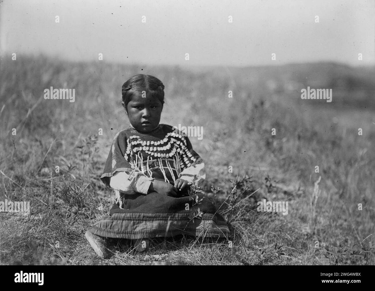 Running Owl's daughter, c1910. Young Piegan girl, full-length portrait, wearing dress decorated with elk teeth, sitting on open ground, facing front. Stock Photo