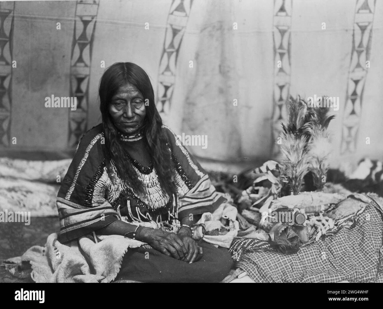Two Kill, c1910. Piegan woman, full-length portrait, seated on blankets inside tipi(?), facing front. Stock Photo