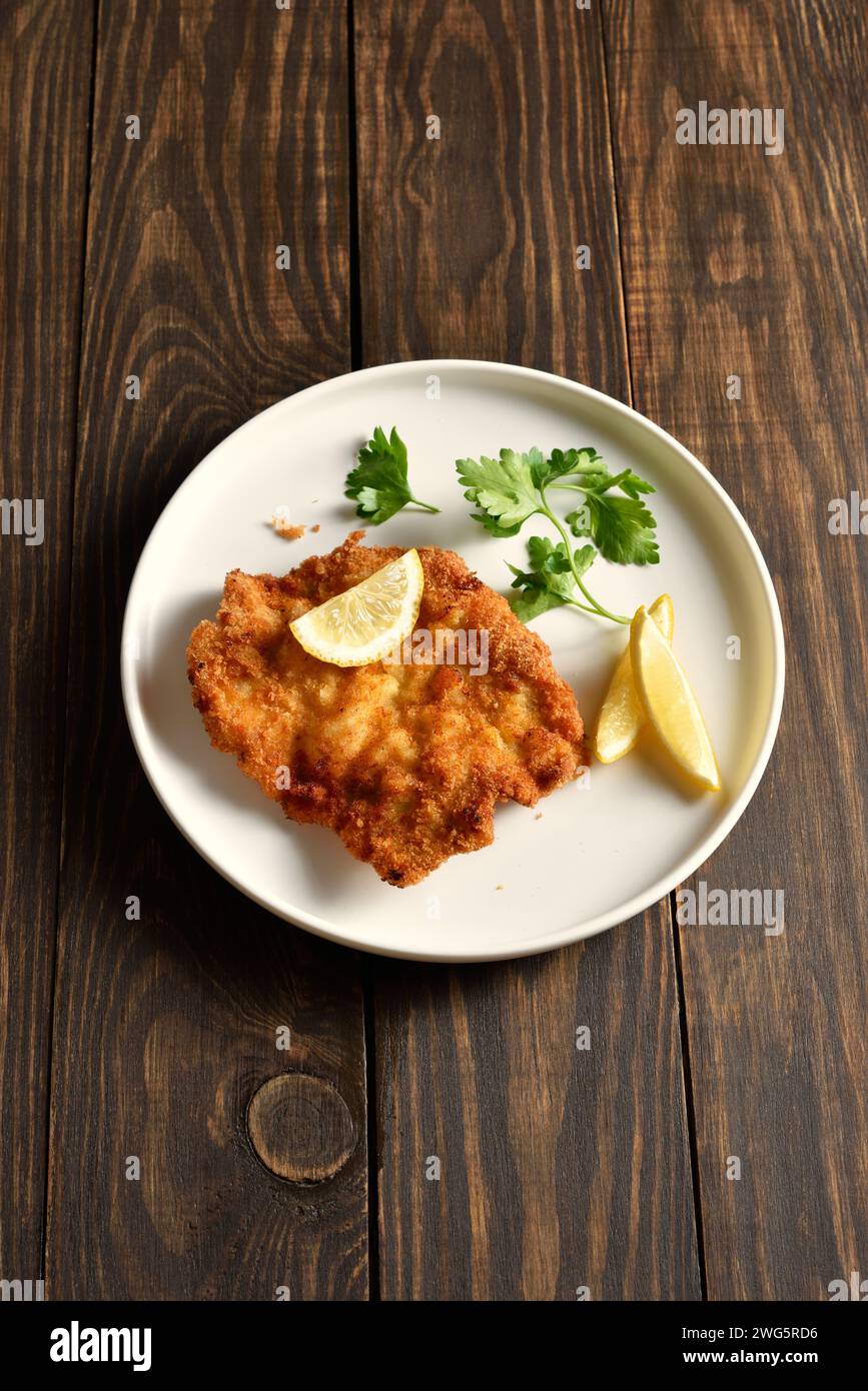 Pork schnitzel served with lemon and leaves of parsley on white plate over rustic wooden background. Country style, close up view Stock Photo