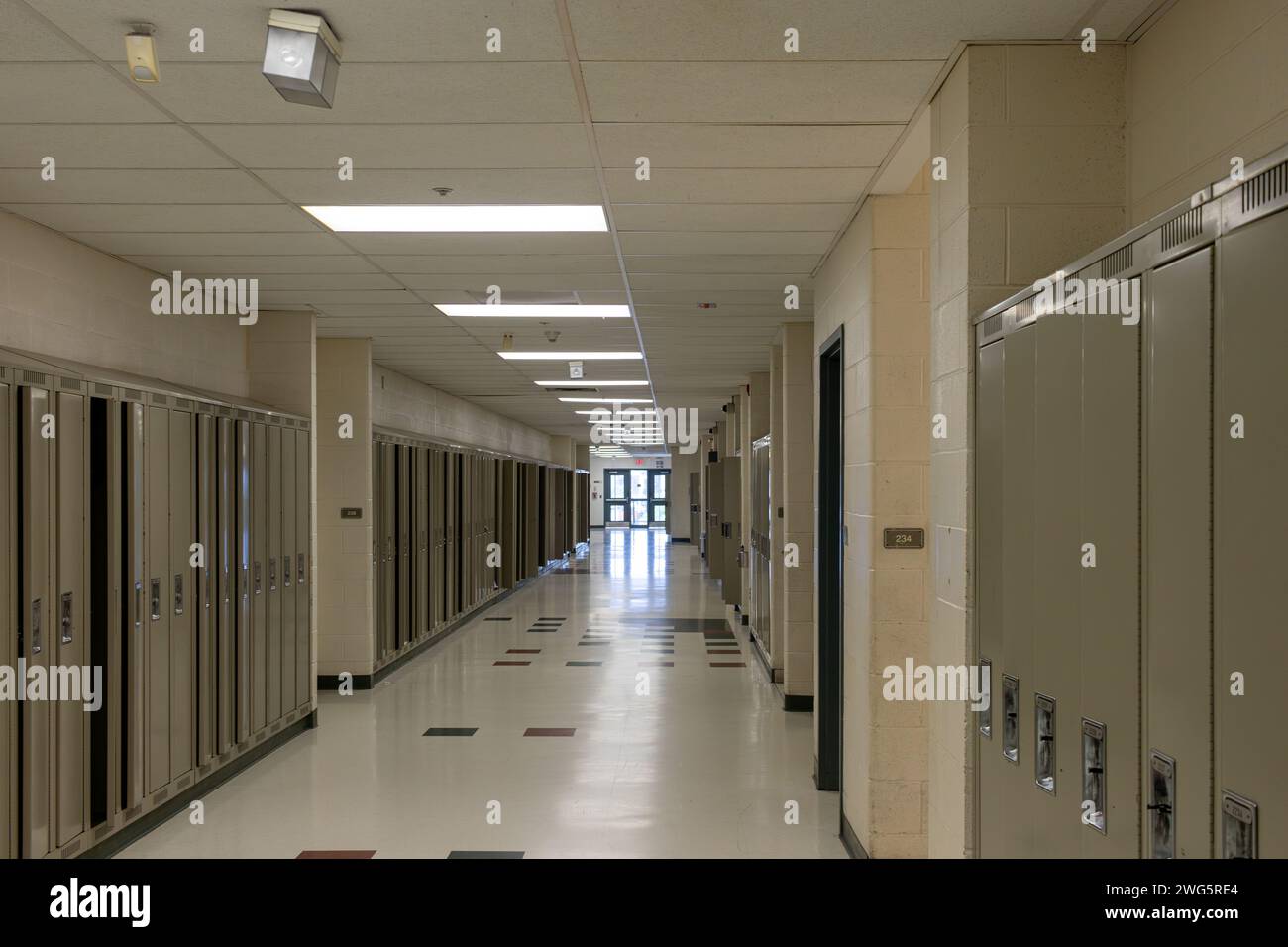 Empty school hallway - lined with closed lockers - beige walls - tiled floor - ceiling lights illuminating space - exit doors at the end. Taken in Tor Stock Photo