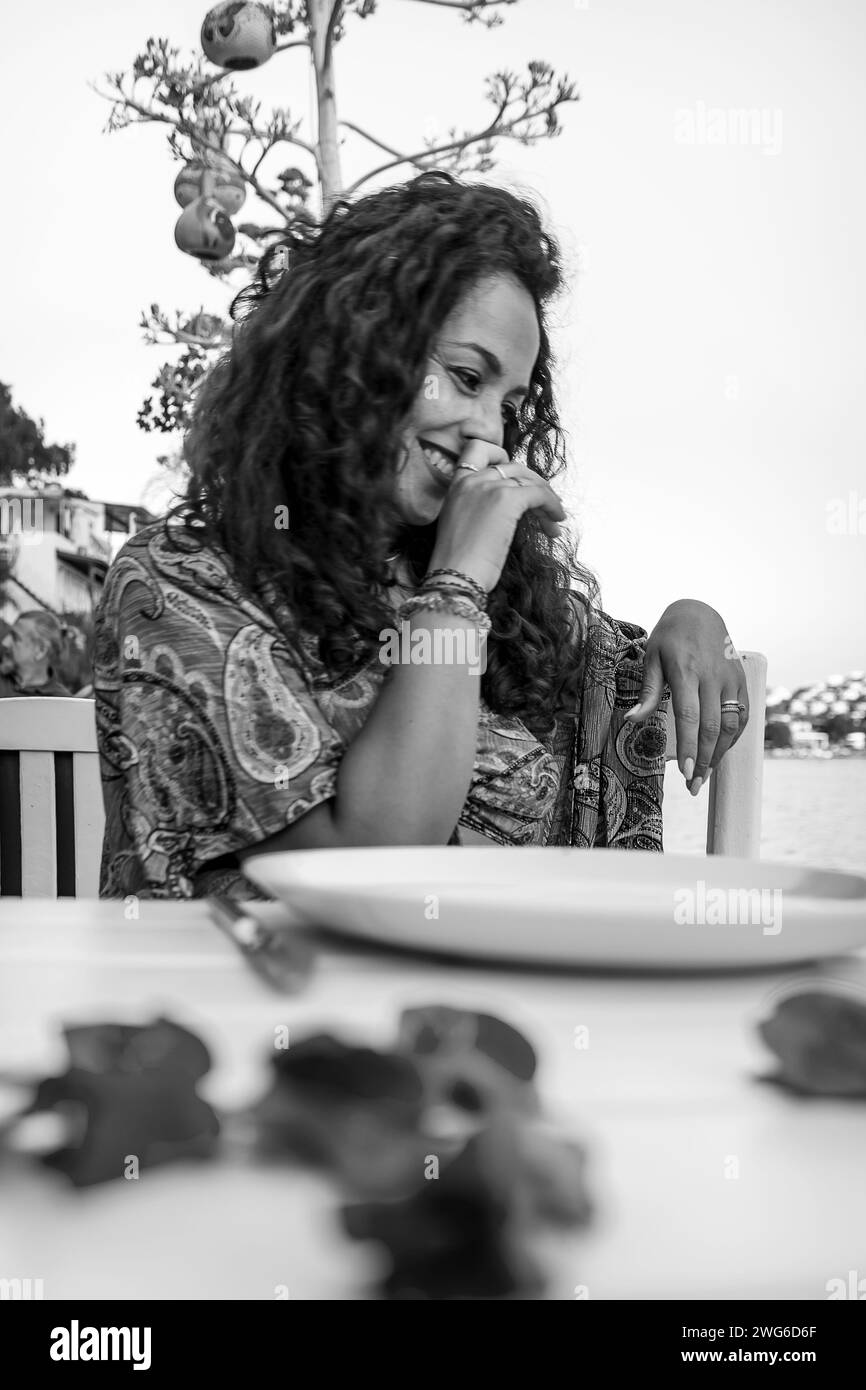 beautiful curly haired mediterranean woman smiles sitting at a restaurant table Stock Photo