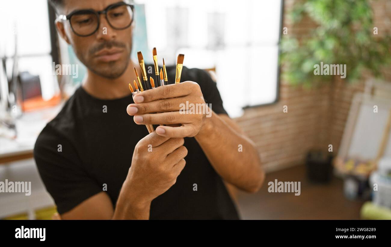 A focused young hispanic man examines paintbrushes in a creative studio setting. Stock Photo