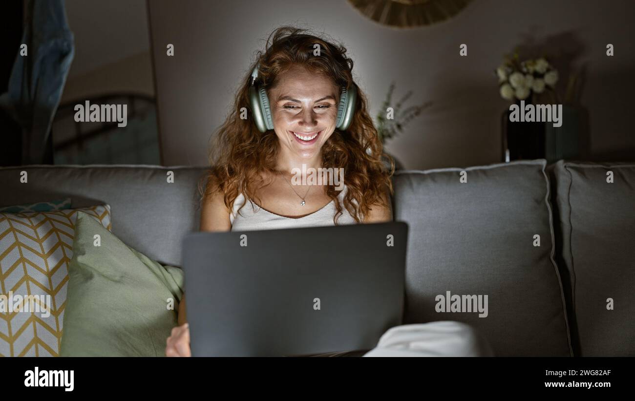 A smiling young woman enjoys music on headphones while using a laptop on the couch in a cozy living room interior. Stock Photo
