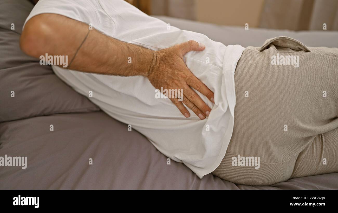 Suffering man, hands touching his injured back, lying on bed in bedroom