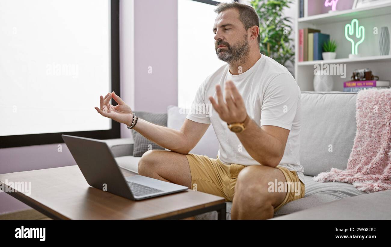 Meditating hispanic man practicing mindfulness at home in living room with laptop Stock Photo
