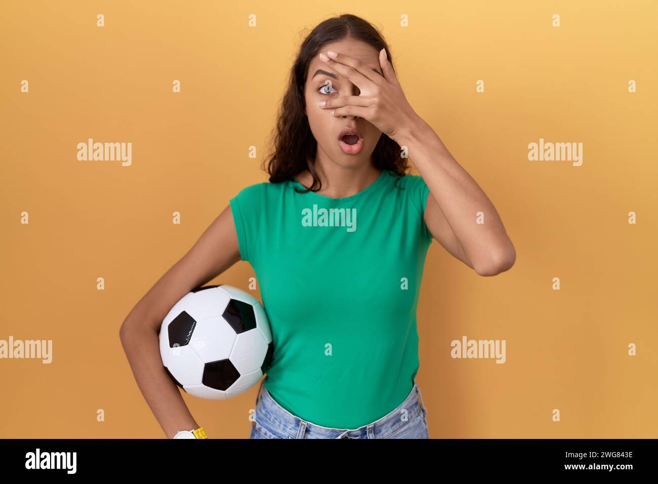 Young hispanic woman holding ball peeking in shock covering face and eyes with hand, looking through fingers with embarrassed expression. Stock Photo