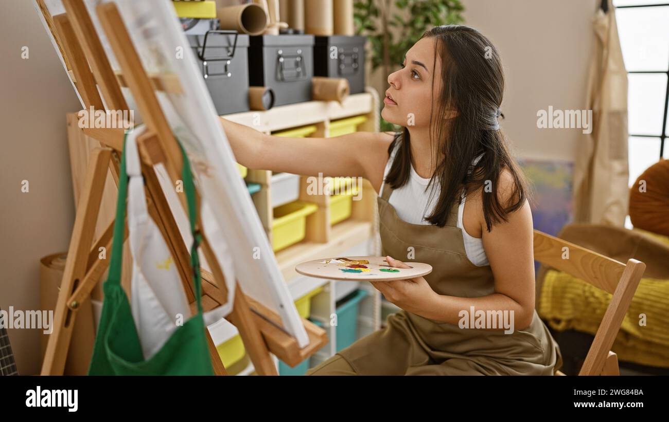Beautiful, focused, young hispanic woman artist sitting on a chair, immersed in creativity, drawing in an art studio, indoors Stock Photo