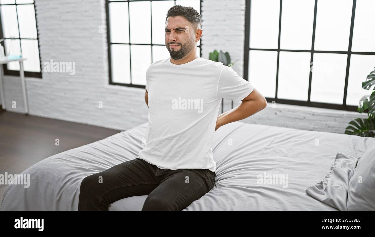 A pained man grimacing and holding his back in a modern bedroom setting Stock Photo